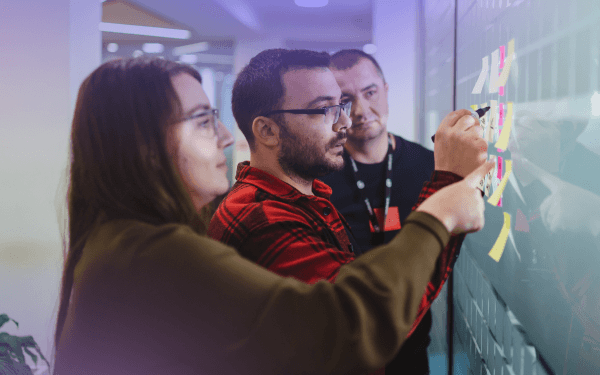 Three people standing in front of a large board covered in sticky notes, engaged in a discussion